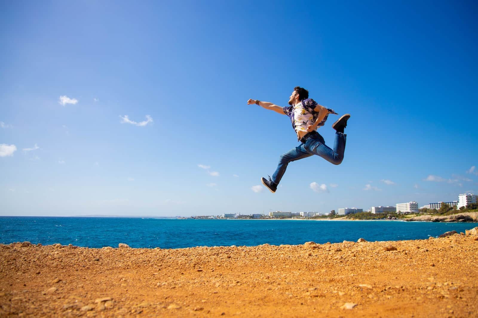Mann in schwarzem Hemd springt tagsüber auf braunem Sand in der Nähe eines Gewässers