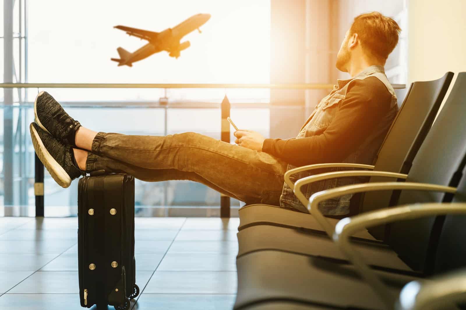Homem sentado em uma cadeira com os pés sobre a bagagem, olhando para um avião